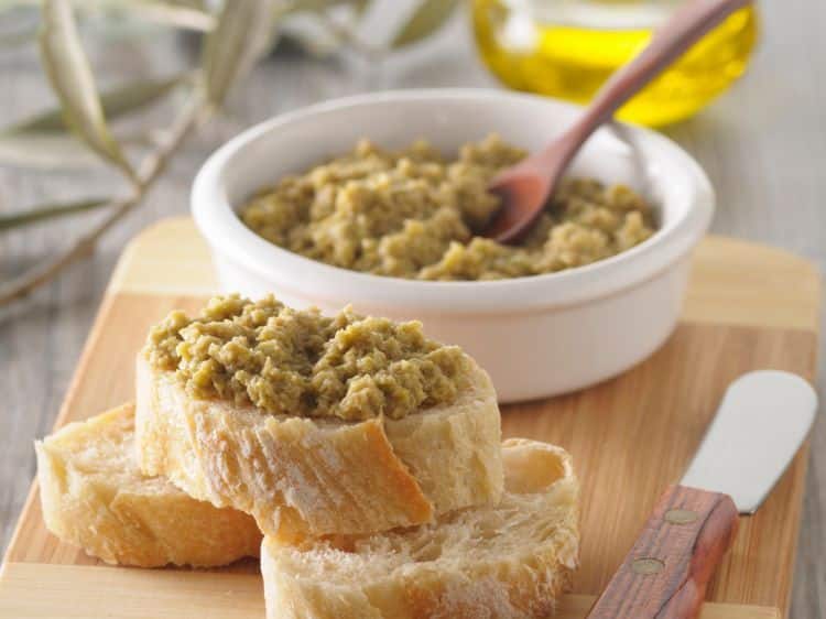 Photo of a small white bowl of tapenade with a wooden spoon, next to a knife and three slices of French baguette bread, on a table with a bottle of olive oil in the background