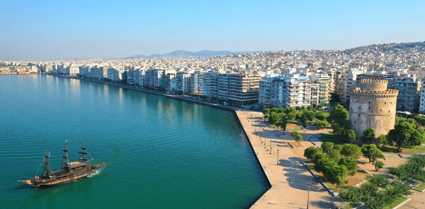 Thessaloniki cityscape with a cruise ship approaching the port, high-rise buildings lining the waterfront, a park with a tower and the city stretching into the distance, all under a clear blue sky with mountains in the background