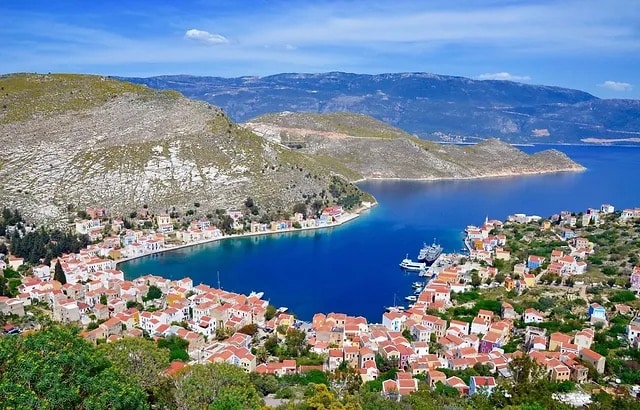 Aerial view of Kastellorizo, a Greek island with whitewashed houses and blue roofs, surrounded by turquoise water, blue sky, mountains and greenery