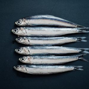 Photo of six raw anchovies on a navy blue background on main page of Growy and Tasty, an Online Greek Market to buy top-quality natural and authentic local products of Greece.