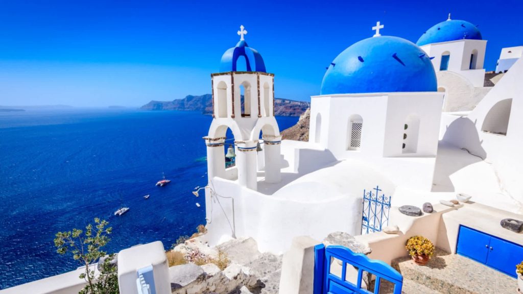 Photo of stunning view of Santorini with whitewashed houses, blue-domed chapel, clear blue sky, sparkling blue Aegean Sea and sailboats