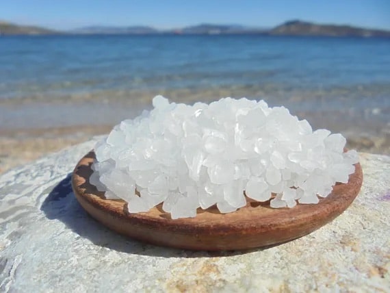 Photo en gros plan de sel de mer grec sur un plateau en bois, situé sur un gros rocher sur une plage avec la mer, les vagues, les montagnes et le ciel bleu en arrière-plan
