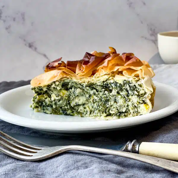 Photo d'une tranche de tarte spanakopita sur une assiette blanche avec une fourchette et un couteau à côté, sur une nappe en tissu avec un fond en marbre. Épicerie Grecque en ligne, avec meilleurs Produits Grecs - Growy and Tasty