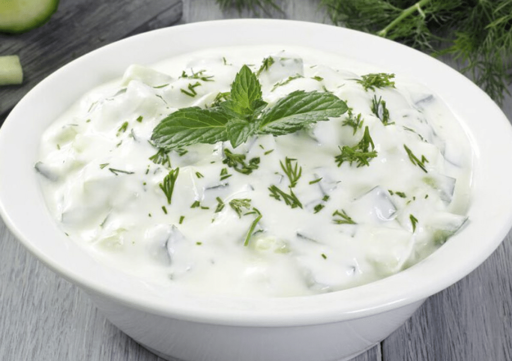 Une photo de tzatziki, une trempette au yaourt grec avec du concombre, de l'ail et des herbes, dans un bol blanc sur une table en bois, entouré d'ingrédients hachés. Boutique de produits grecs en ligne - Growy and Tasty