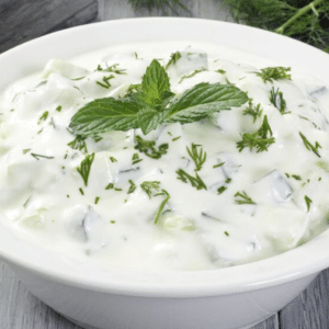 A photo of tzatziki, a Greek yogurt dip with cucumber, garlic and herbs, in a white bowl on a wooden table, surrounded by chopped ingredients by Growy and Tasty | Greek Online Shop Where You Can Buy Local products of Greece.