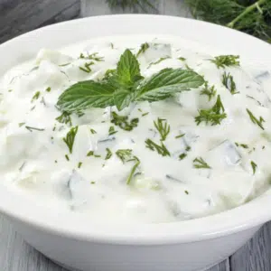 Une photo de tzatziki, une trempette au yaourt grec avec du concombre, de l'ail et des herbes, dans un bol blanc sur une table en bois, entouré d'ingrédients hachés. Boutique de produits grecs en ligne - Growy and Tasty