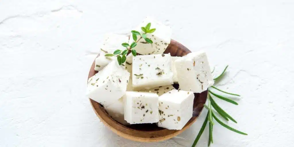 Greek feta cheese cubes in wooden bowl with rosemary and herbs on white background