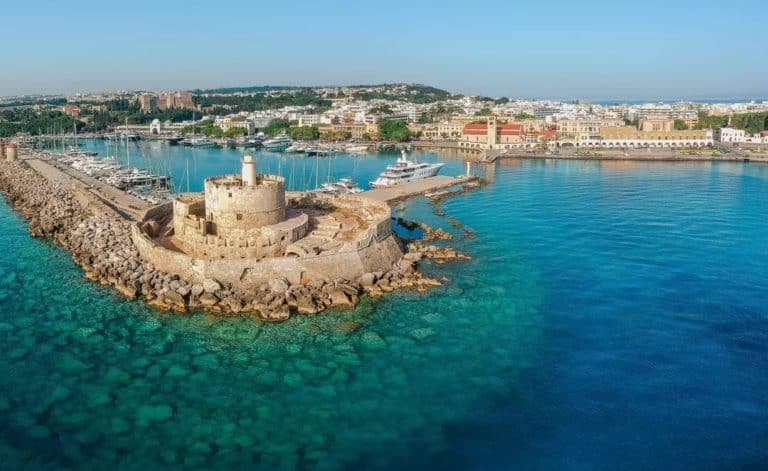 Vue panoramique du port de Rhodes en Grèce, mettant en vedette le célèbre phare, l'entrée du port, les bateaux de plaisance, la ville et le château, l'eau turquoise et le ciel bleu