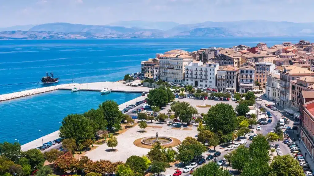 Πανοραμική θέα της Κέρκυρας με θάλασσα, βουνά, μπλε ουρανό, σπίτια, πολυσύχναστη πλατεία, αυτοκίνητα και βάρκες στο λιμάνι