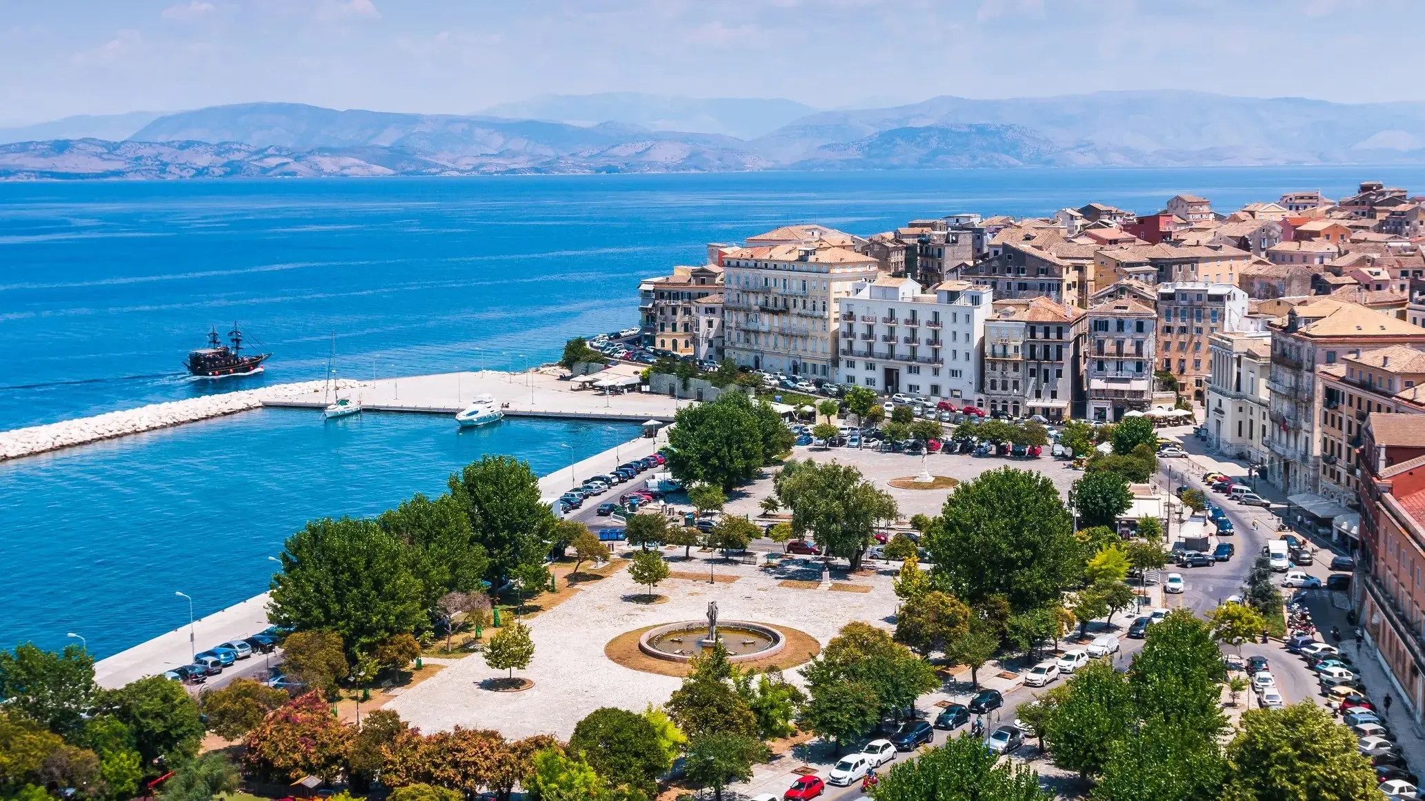 Vue panoramique de Corfou avec mer, montagnes, ciel bleu, habitations, place animée, voitures et bateaux dans le port