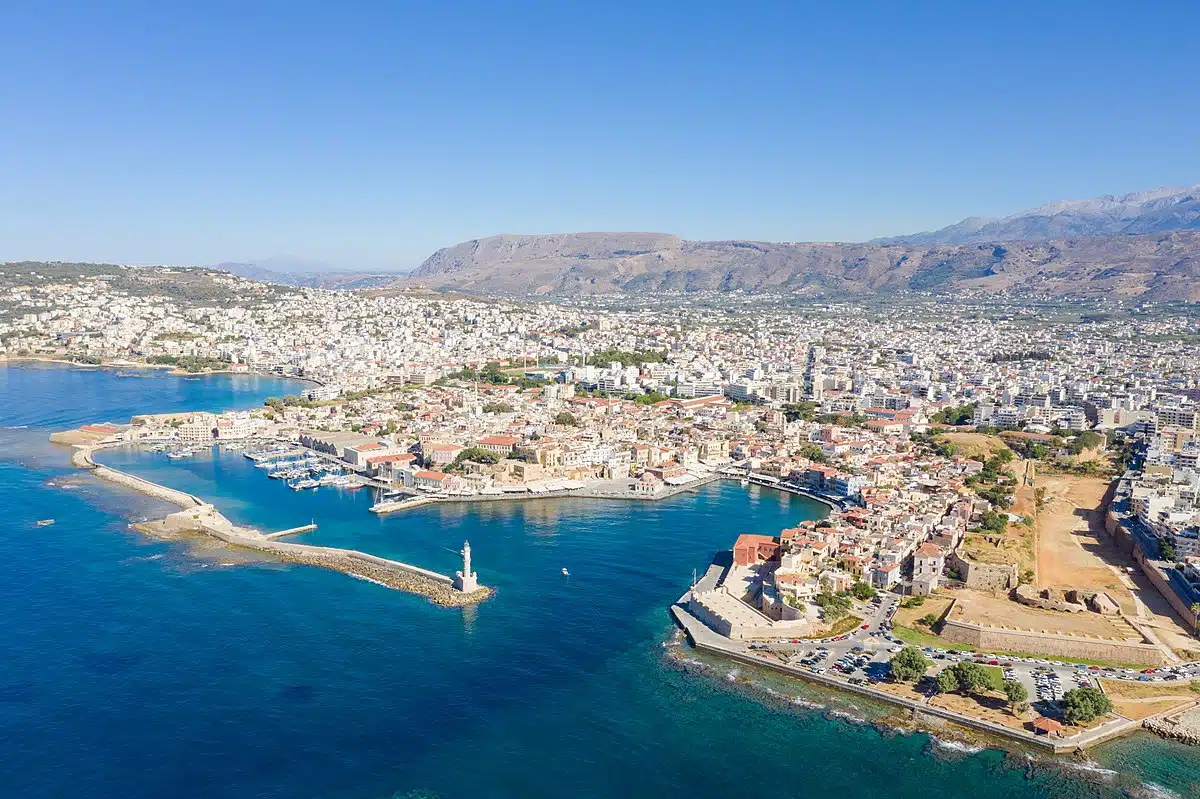 Photo of the port of Chania in Crete, showing the Venetian port with its moored boats, surrounding buildings, lighthouse, blue sky and sea