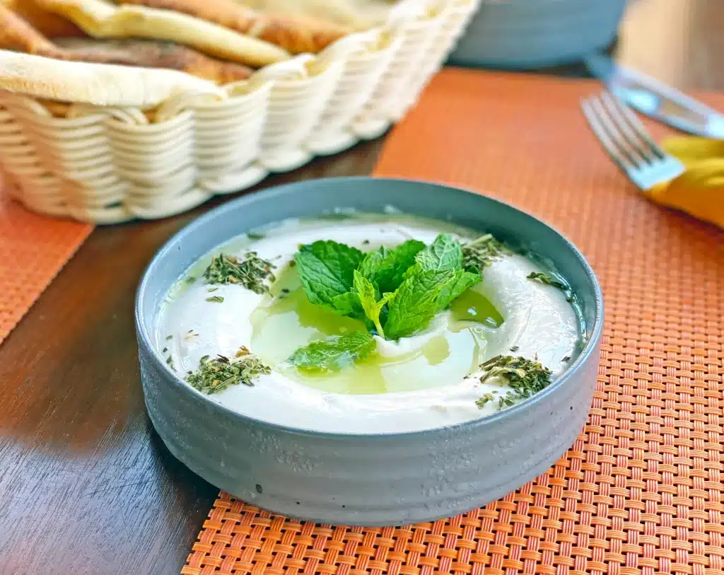 Fresh tzatziki with mint on wooden table with basket of Greek breads, herbs, fork and orange placemats