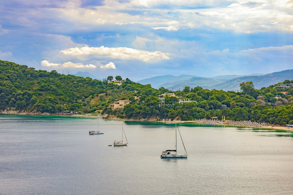 Baie de Skiathos en Grèce avec bateaux à voile ancrés, plage bordée de pins et collines verdoyantes Doit décrire l’image de façon claire et intégrer naturellement les mots-clés : Skiathos, Grèce, plage, bateaux.