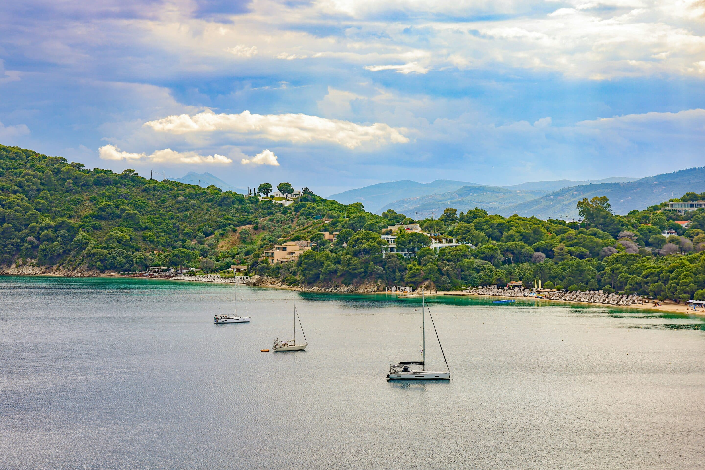 Baie de Skiathos en Grèce avec bateaux à voile ancrés, plage bordée de pins et collines verdoyantes Doit décrire l’image de façon claire et intégrer naturellement les mots-clés : Skiathos, Grèce, plage, bateaux.