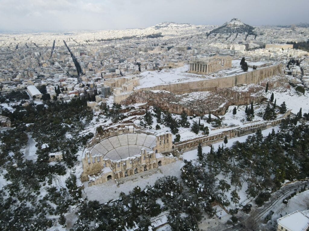 Vue aérienne de l’Odéon d’Hérode Atticus et de l’Acropole d’Athènes sous la neige, avec la ville moderne en arrière-plan.