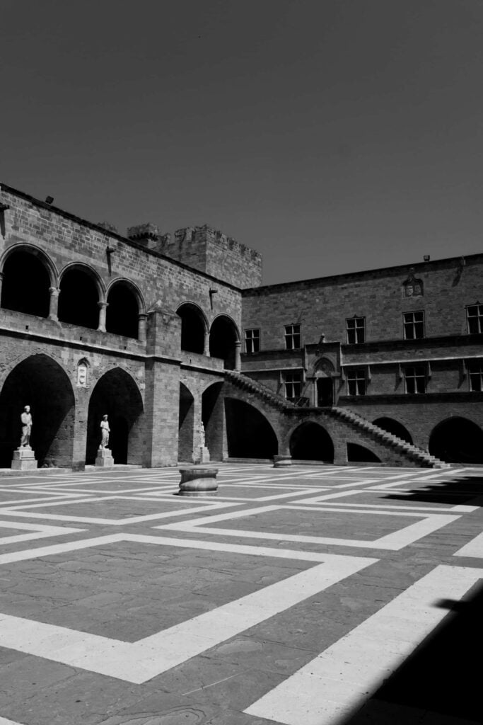 Vue en noir et blanc du cour intérieure du Palais des Grands Maîtres à Rhodes, avec des arcades et des statues.