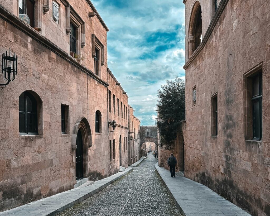 Rue pavée dans la vieille ville de Rhodes, avec des bâtiments médiévaux et une vue partielle du Palais des Grands Maîtres.