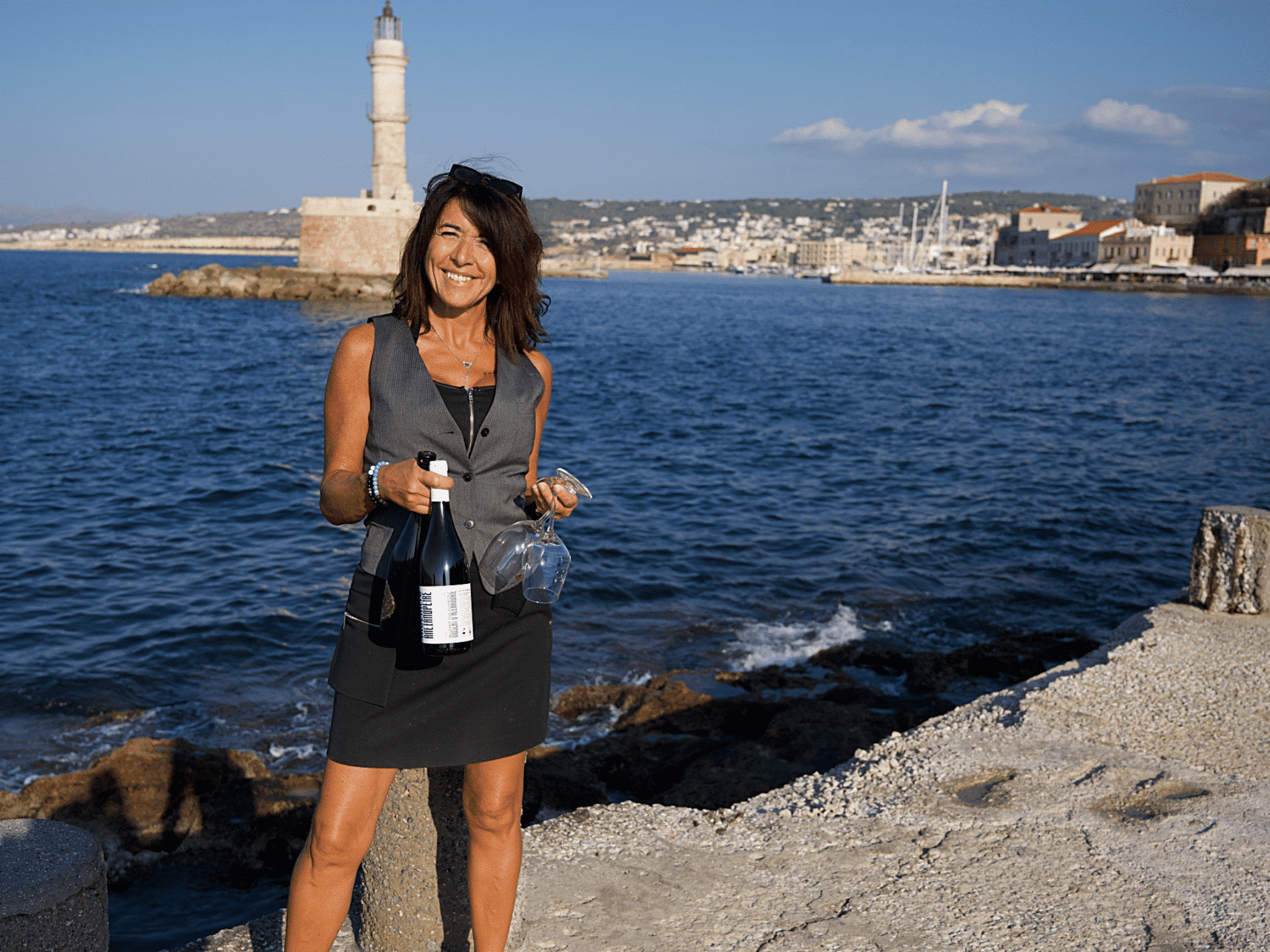 Myriam présentant une bouteille de vin et des verres devant le phare du vieux port de Chania, promotion de la dégustation de vin à la Cave de Platonie.