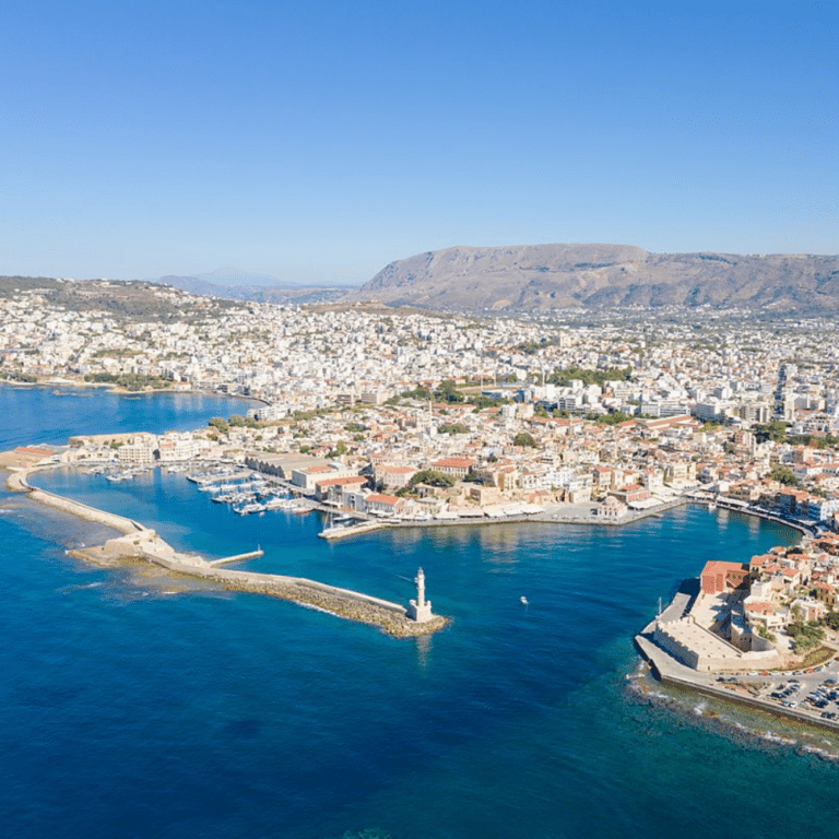 Photo du port de La Canée en Crète, montrant le port vénitien avec ses bateaux amarrés, les bâtiments environnants, le phare, le ciel bleu et la mer. Les régions en Grèce.